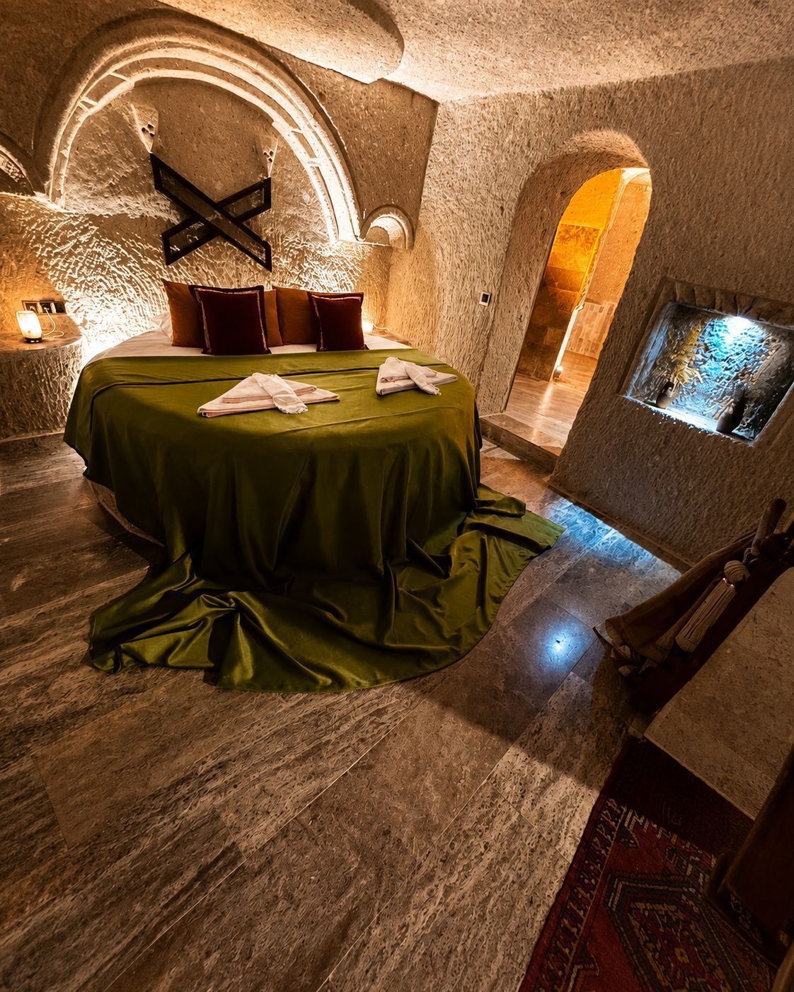 A circular bed with a green bedspread, a wooden bench, and a stone wall with a window.