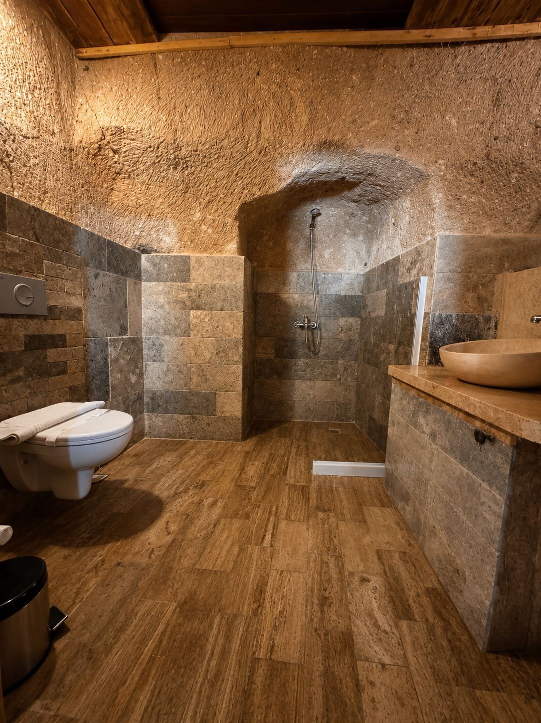 Un baño con pared de piedra, suelo de madera, inodoro, ducha y lavamanos.