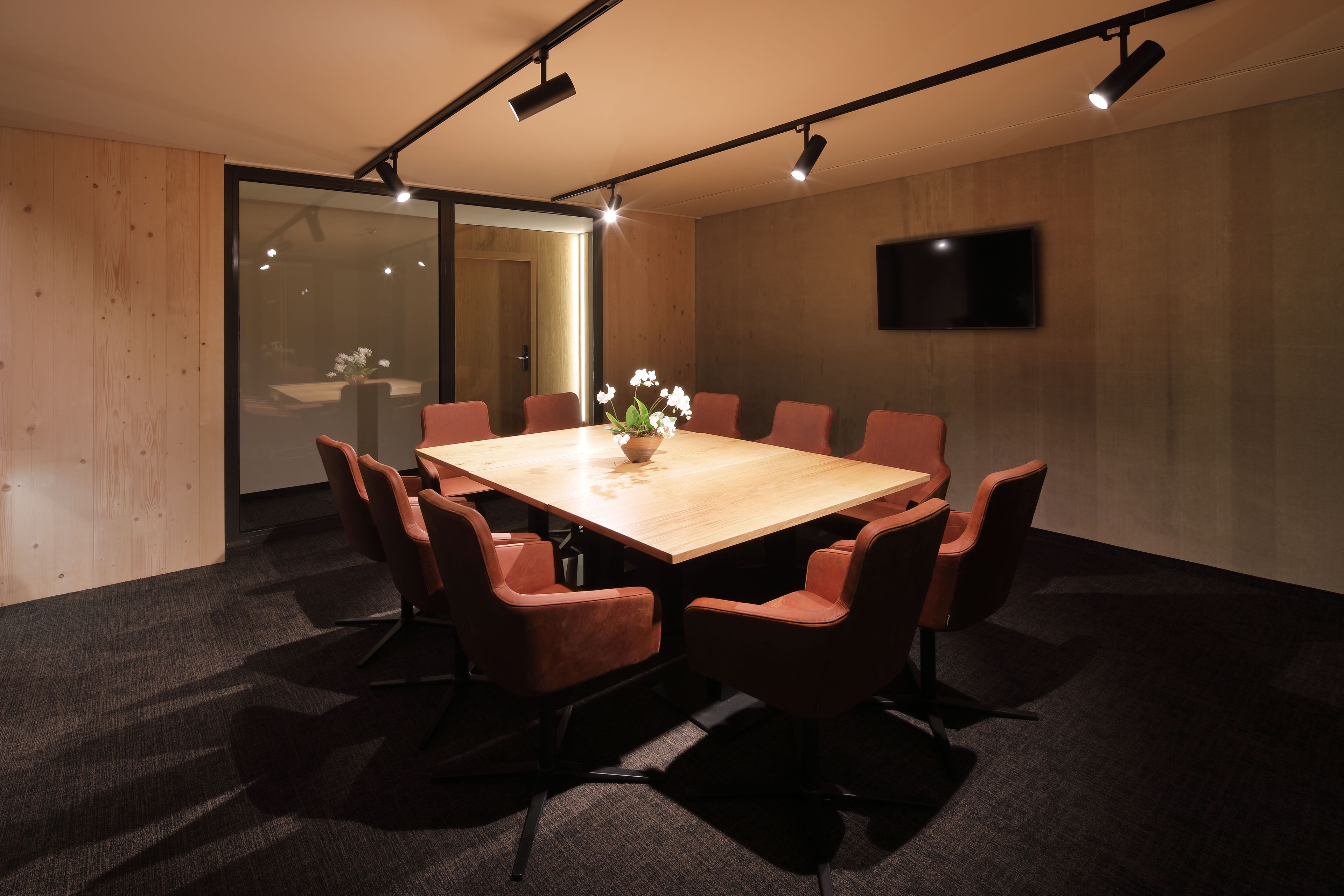 A conference room with a wooden table, eight brown chairs, a vase with flowers, and a flat-screen TV on the wall.