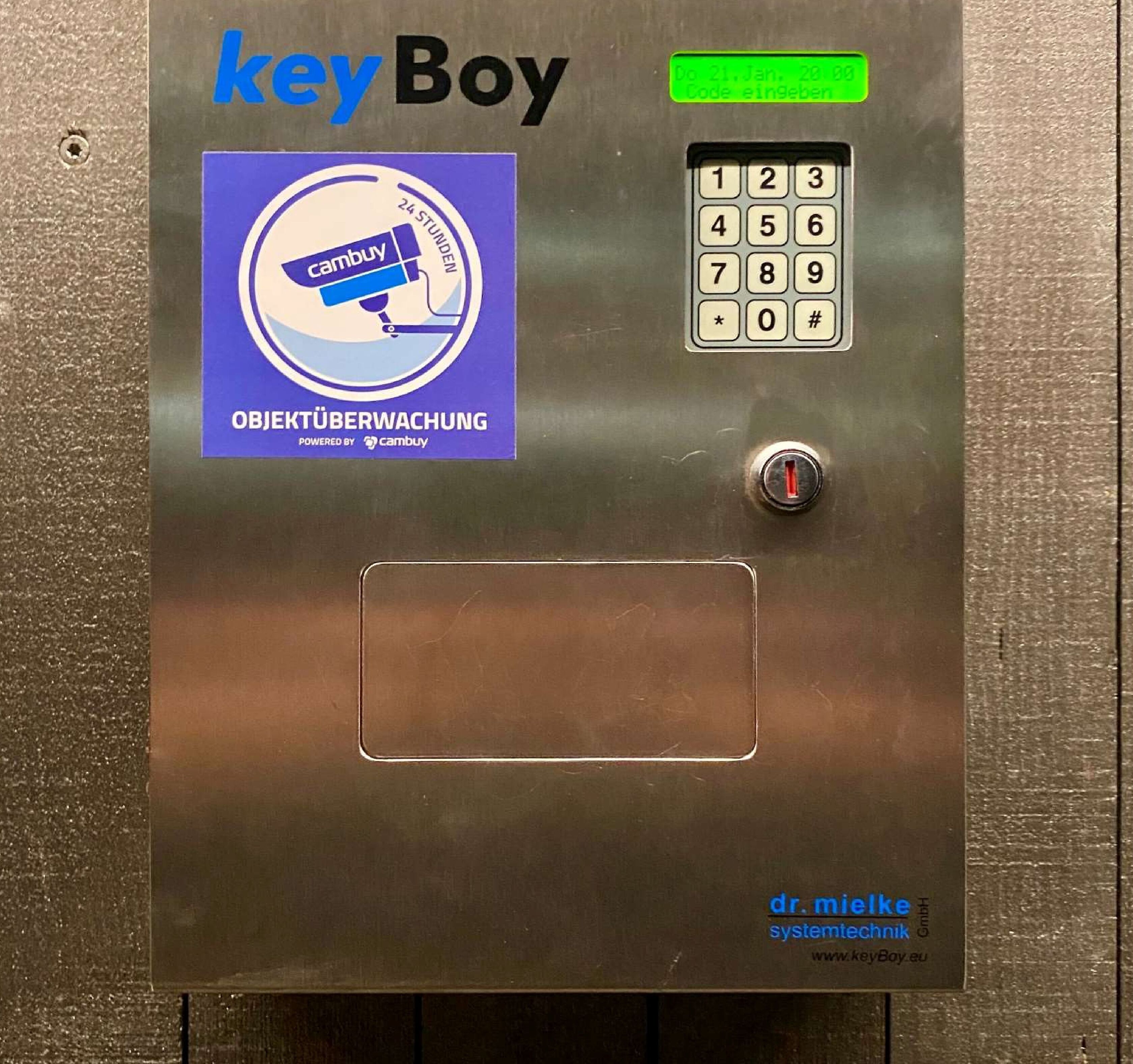 Ein Bedienfeld einer Aufzugskonsole mit Tastatur, einem grünen Knopf und der Aufschrift 'keyBoy' und 'OBJECTIVE WATCHING'.
