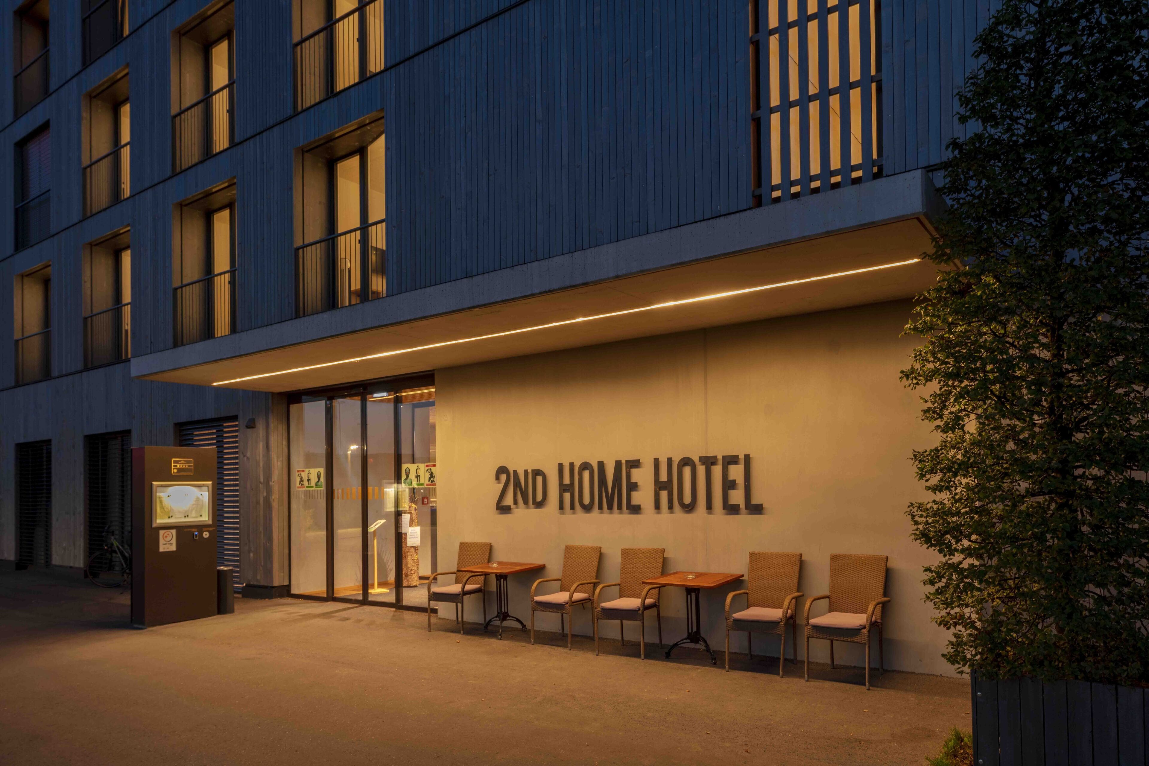 Entrada de hotel con un letrero que dice "2ND HOME HOTEL", asientos exteriores con sillas y fachada de edificio con ventanas y una puerta.