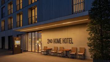 Entrada de hotel con un letrero que dice "2ND HOME HOTEL", asientos exteriores con sillas y fachada de edificio con ventanas y una puerta.