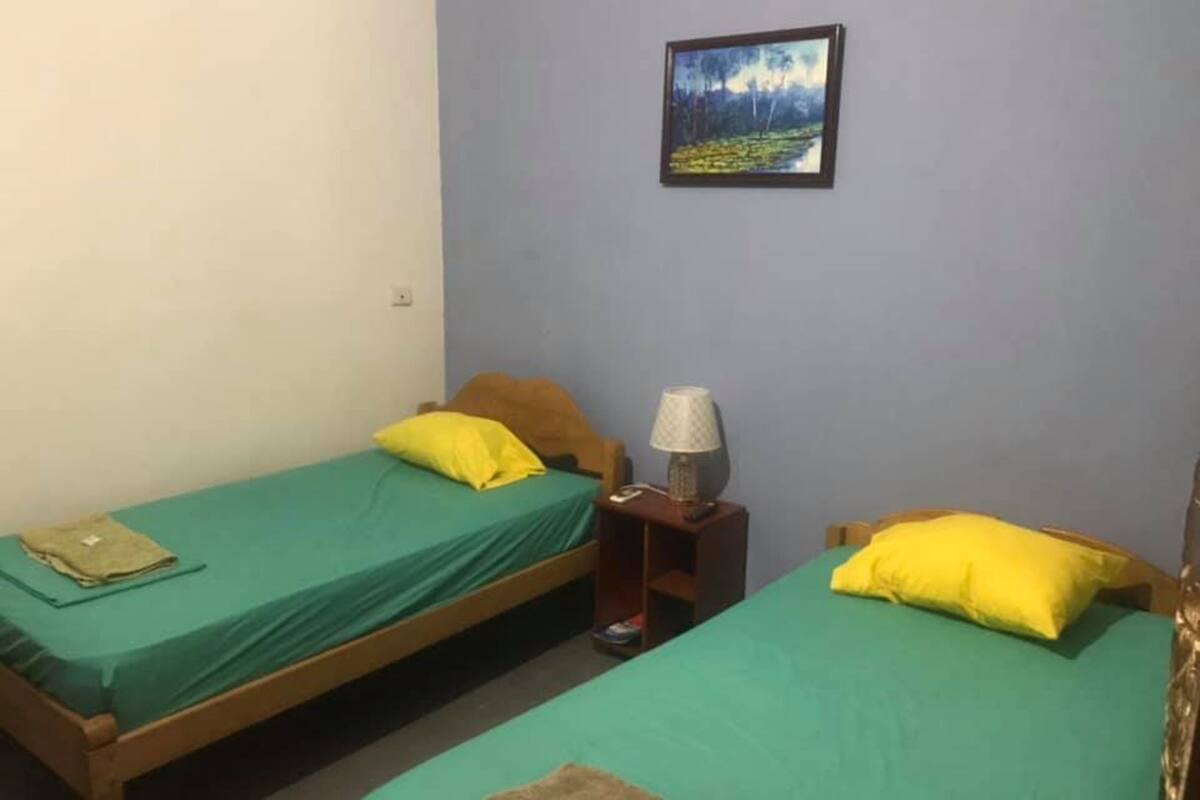 A room with two single beds, each with a green mattress and a yellow pillow. There is a small table with a lamp between the beds.