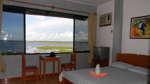 A hotel room with a bed, a desk, a chair, a television, and a window with a view of a body of water and greenery. - Amazon Dream Hostel (Iquitos)
