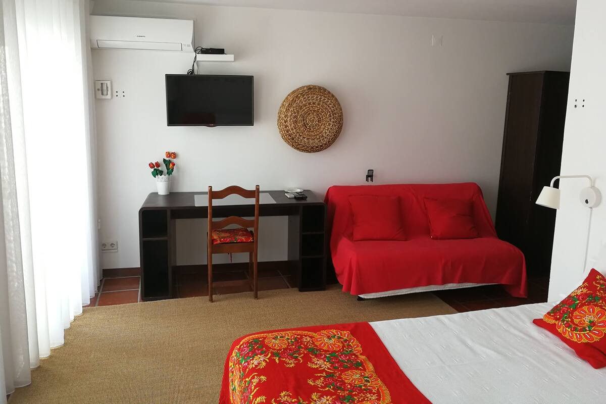 Een hotelkamer met een bed, een rode bank, een bureau met een computer, een televisie en een luchtverkoeler aan de muur.