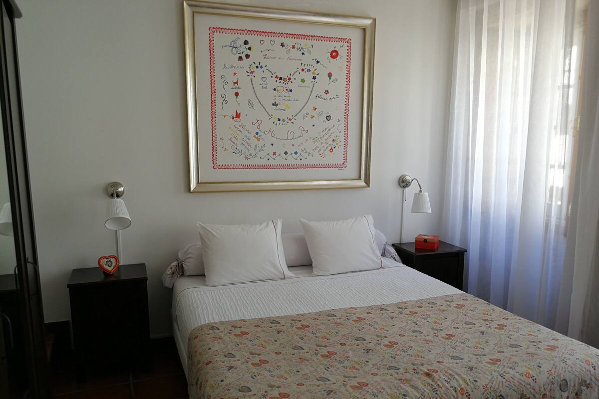 Een netjes ingerichte slaapkamer met een bed, twee nachtkastjes, een lijst met kunstwerk en een raam met gordijnen.