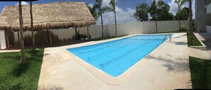 Una piscina con una estructura de techo de paja, rodeada por una terraza de concreto y palmeras.