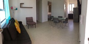 Una sala de estar con un sofá, una mesa de comedor con sillas, una zona de cocina y un acceso a otra habitación.