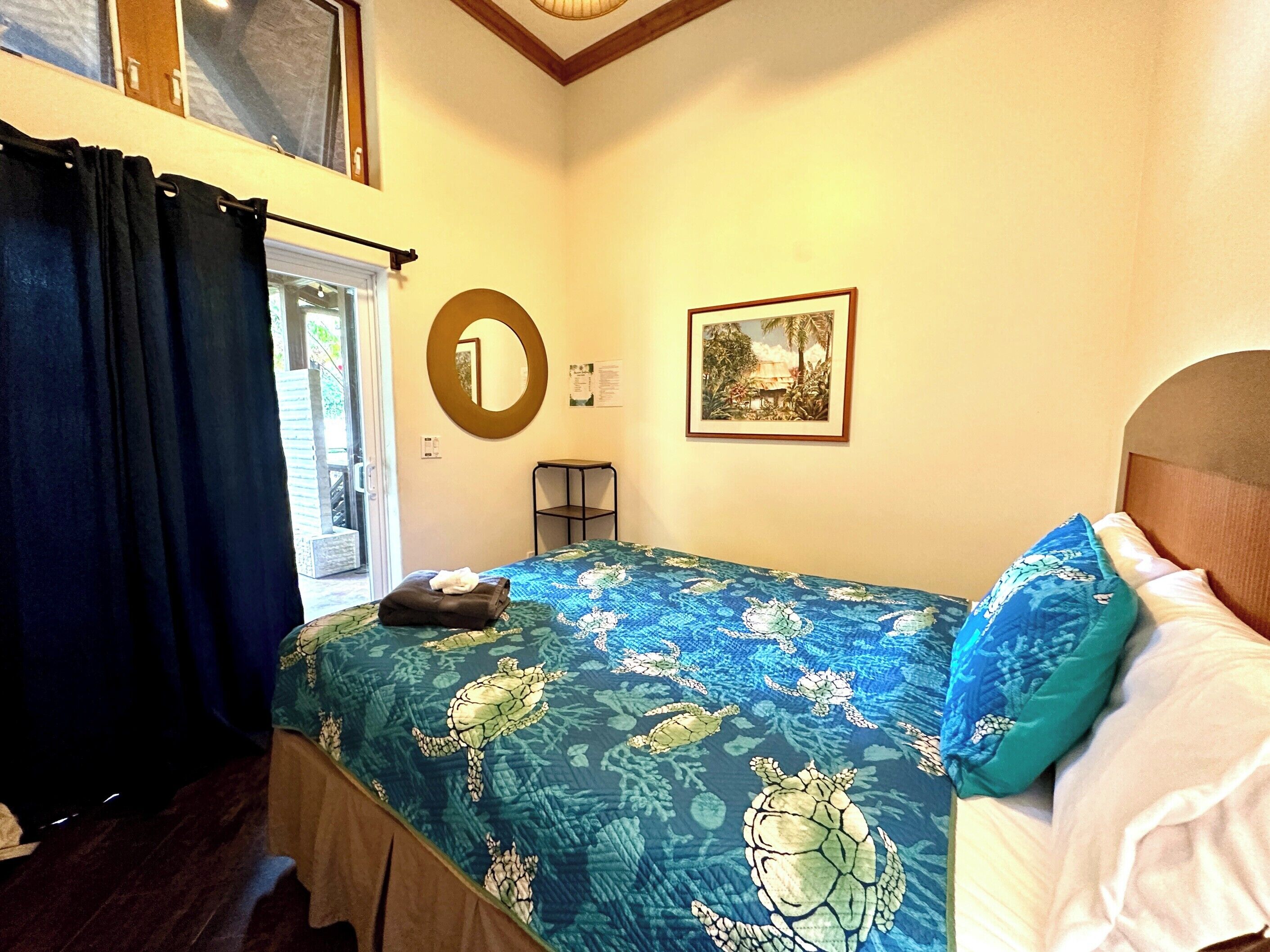Une chambre à coucher avec un lit, une fenêtre avec des rideaux, une petite table, un miroir et un tableau encadré au mur.