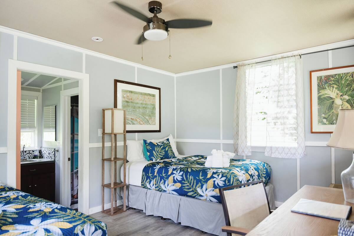 A bedroom with two beds, a ceiling fan, a window with sheer curtains, and a framed picture on the wall.