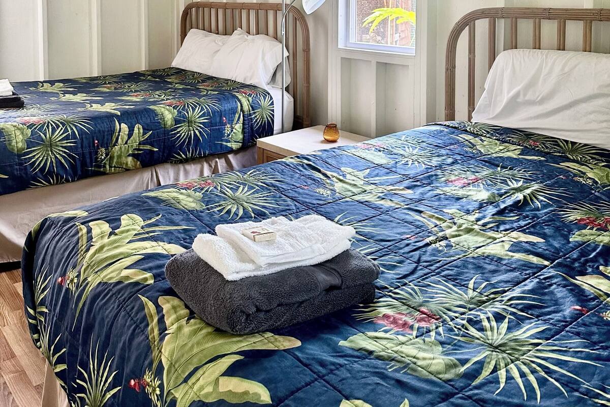 A bedroom with two single beds, a bedside table with a lamp, and a window with a view of greenery.