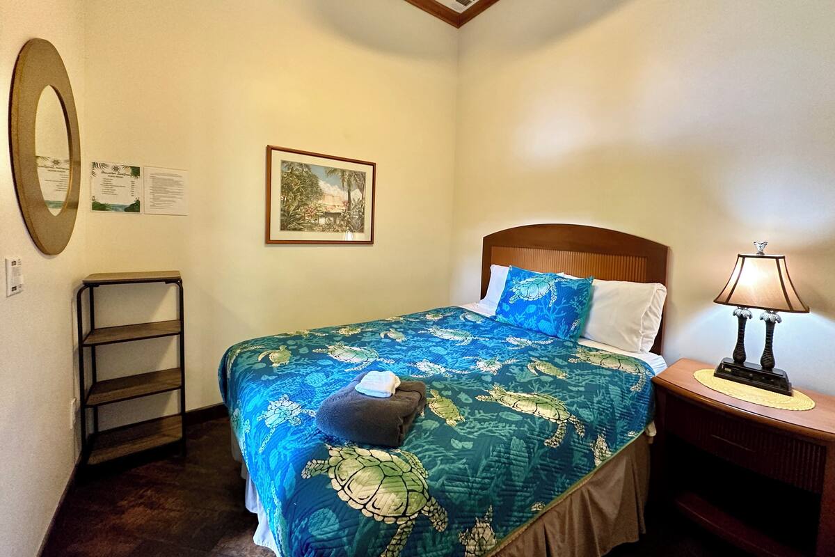 A neatly arranged hotel room with a bed, a bedside lamp, a framed picture on the wall, and a small shelf.