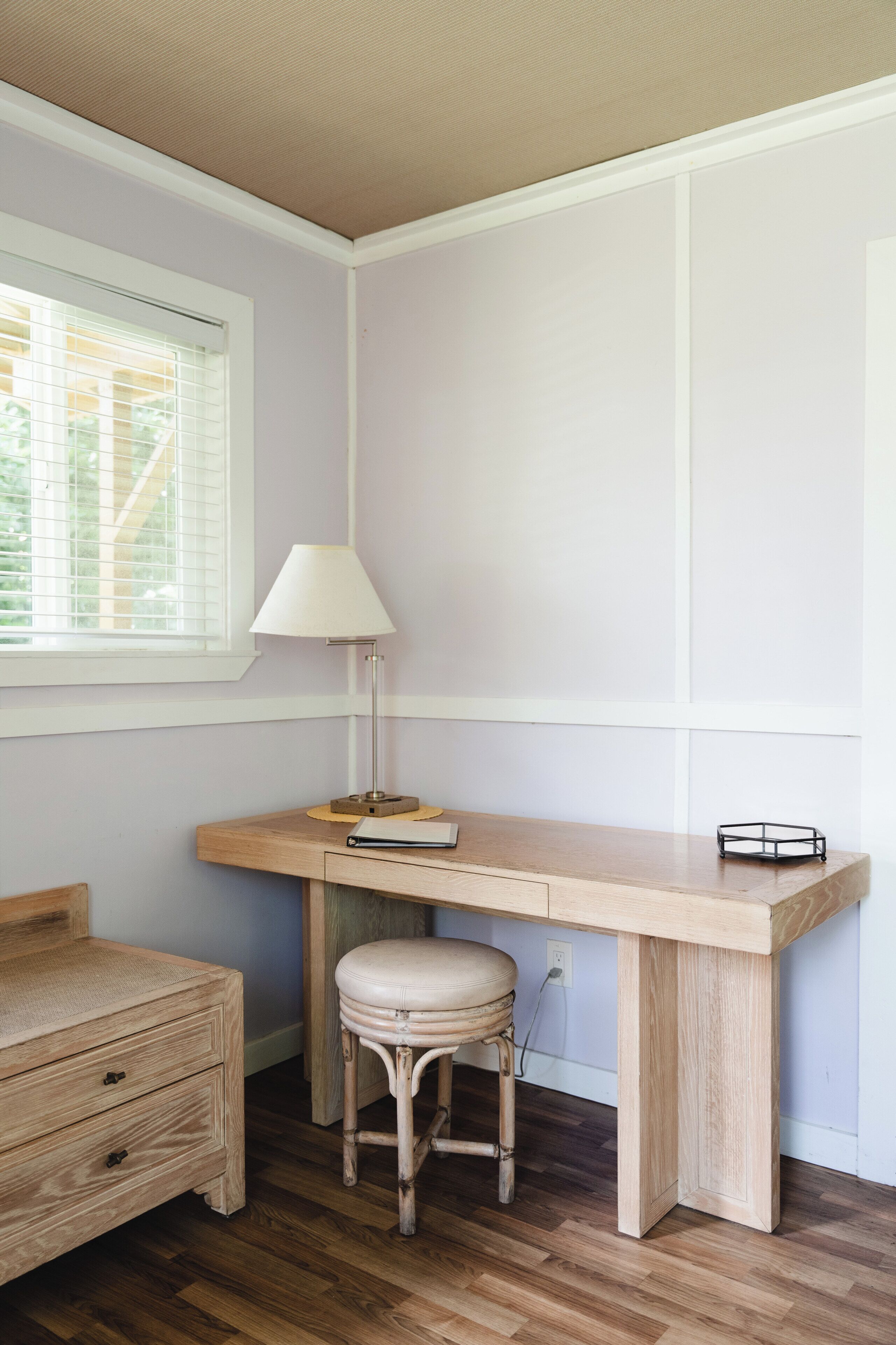 Un espacio de estudio con un escritorio de madera, una lámpara blanca, un taburete de mimbre y una ventana con persianas.