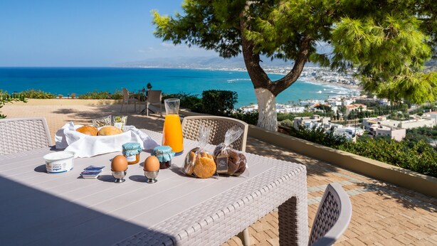 A breakfast table with food and drinks, overlooking a coastal view with a tree and buildings.