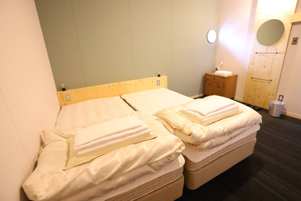 A double bed with two pillows and a wooden headboard, a small wooden nightstand with a mirror and a towel, and a metal trash bin.