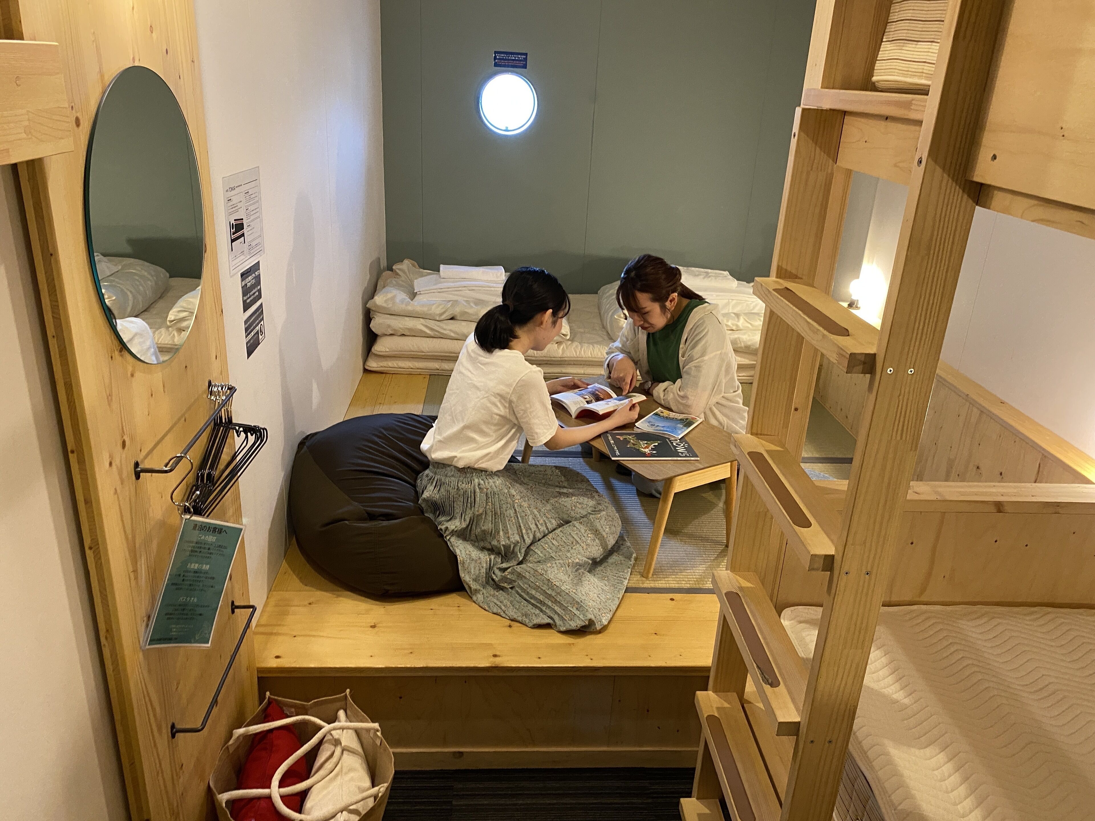 A cozy room with a bed, a small table, and a mirror. There are two people sitting at the table, one reading a book.