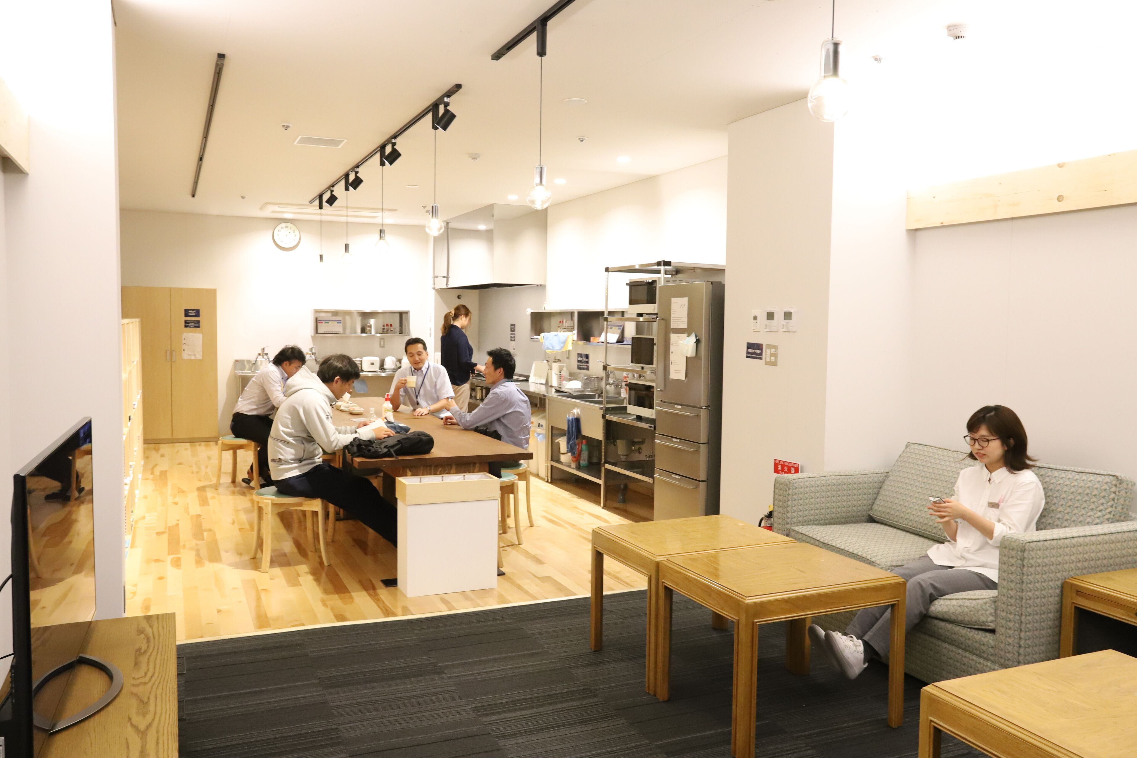 Un área de cocina moderna con suelo de madera, electrodomésticos de acero inoxidable y un grupo de personas conversando alrededor de una mesa.