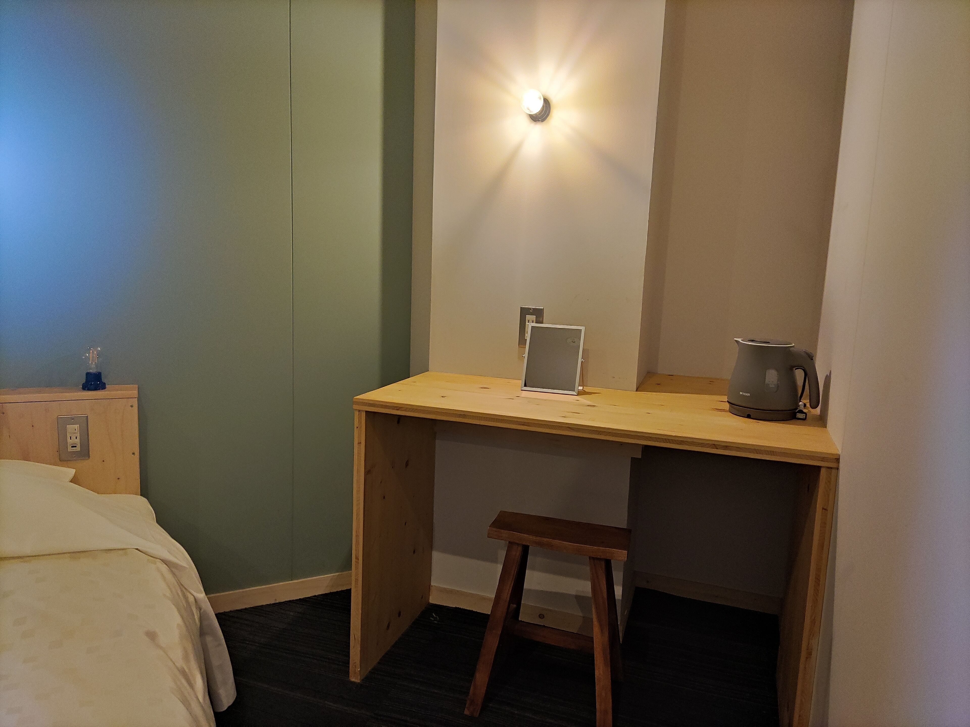 A small, neatly organized room with a wooden desk, a stool, a bed, and a wall-mounted light.