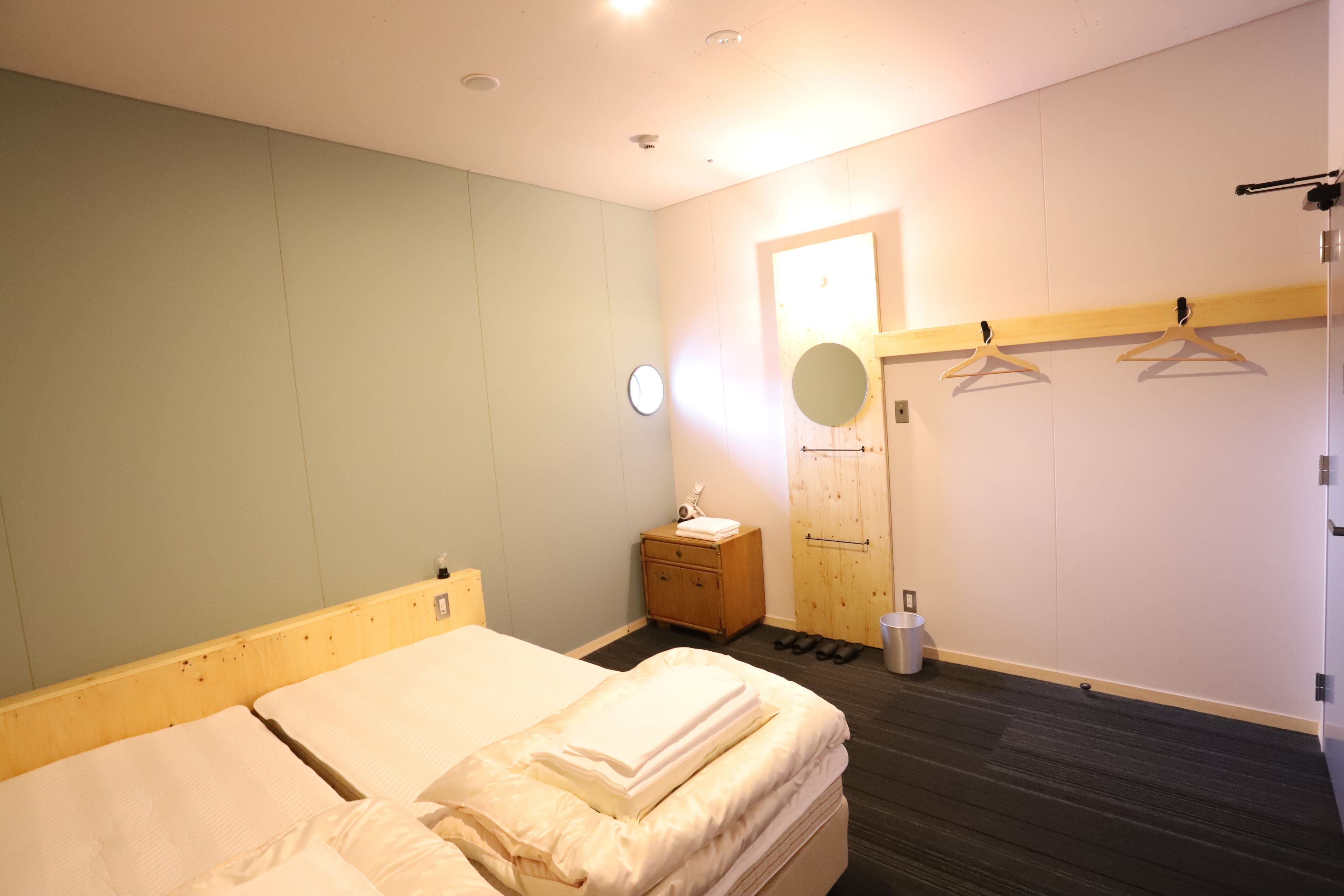 A hotel room with a bed, a wooden nightstand, a mirror, a coat rack, and a door with a light.