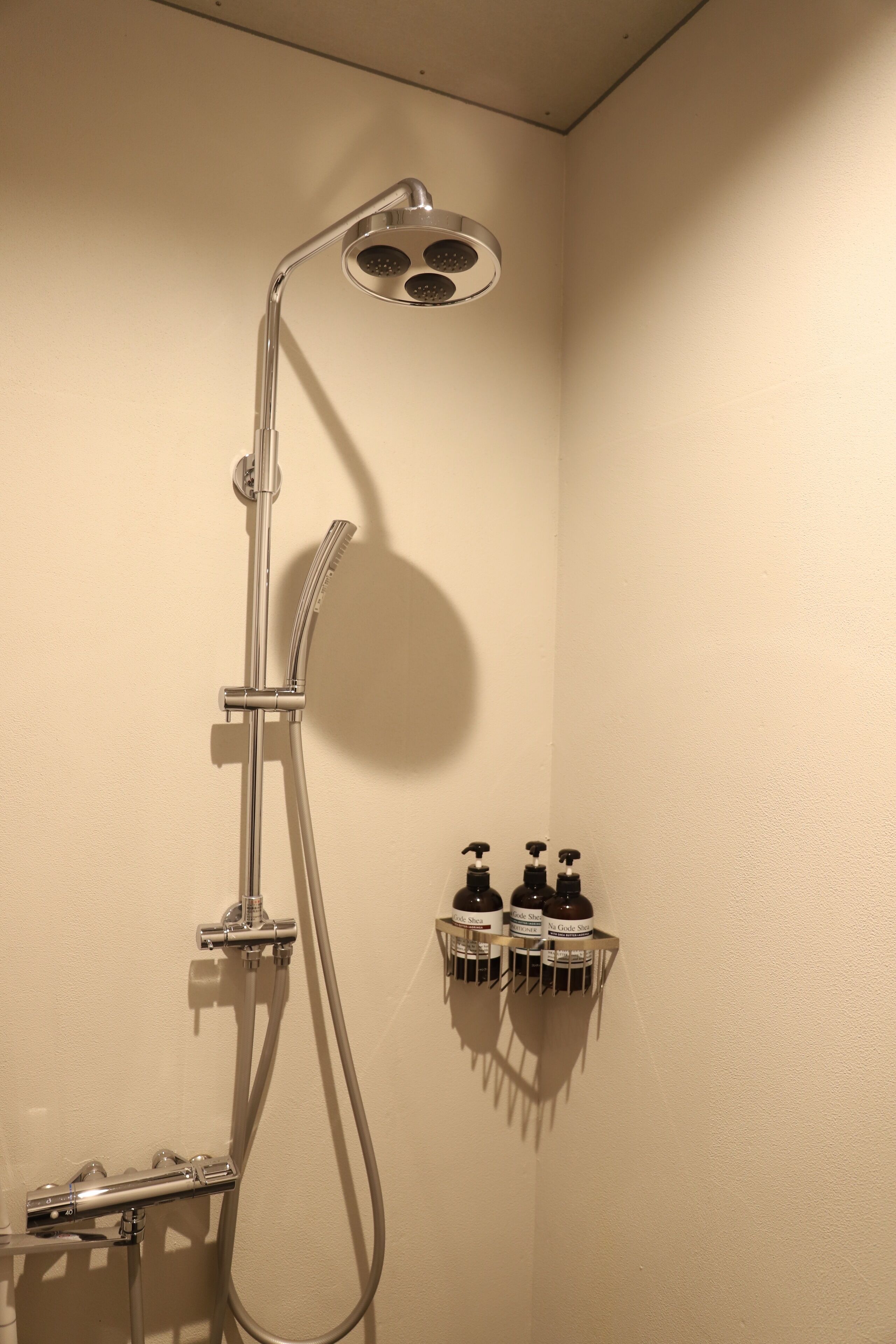 A shower with a handheld showerhead and a wall-mounted faucet. There are three bottles of shampoo on a shelf below.
