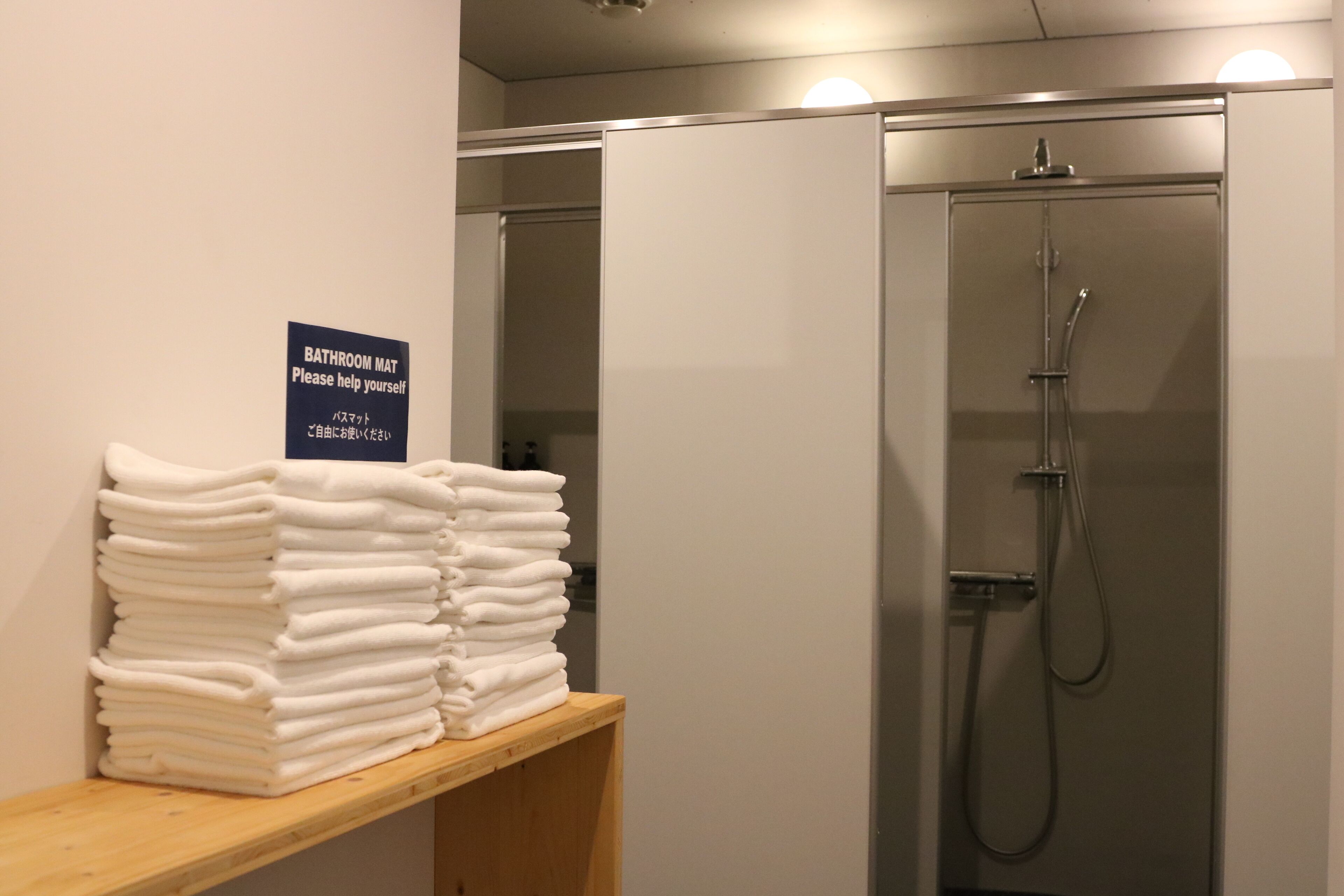 A stack of white towels on a wooden shelf with a sign that reads "BATHROOM MAT Please help yourself."