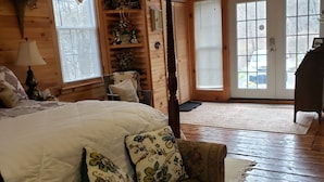 Ein gemütliches Zimmer mit einem großen Bett, Holzboden, einem Sessel mit floralen Kissen, einer Lampe und einem Fenster mit Jalousien.