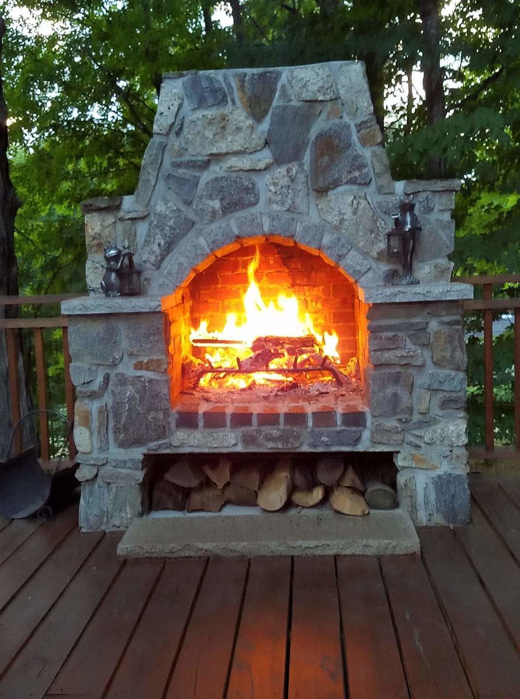 A stone-built outdoor fireplace with a fire burning inside, surrounded by a wooden deck.