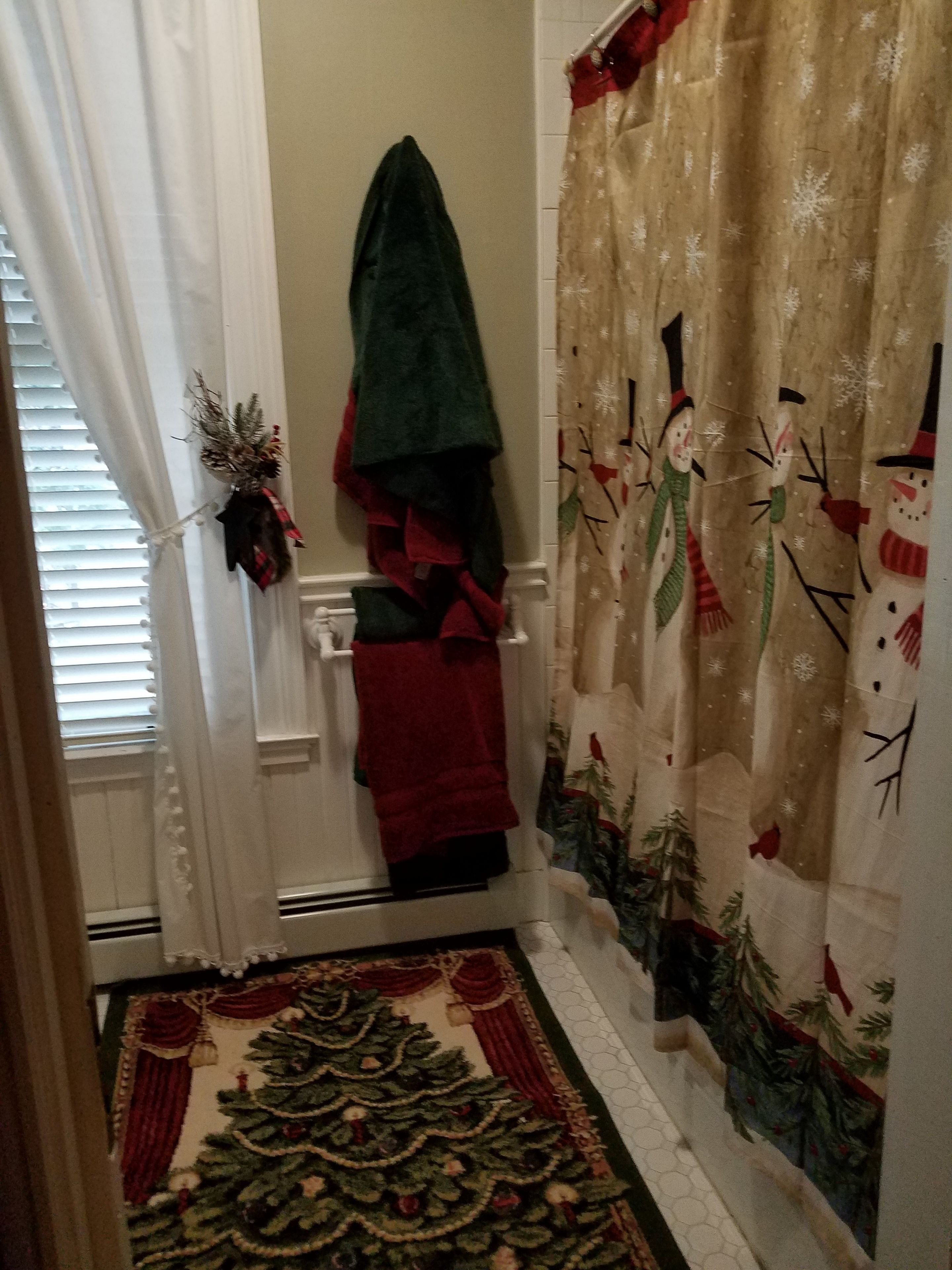 Une salle de bain avec un rideau de douche orné de motifs de bonhommes de neige et d’un sapin de Noël, un porte-serviettes avec une serviette verte, et un tapis à motifs de sapin de Noël sur le sol.