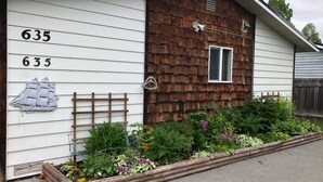 Una casa con el número 635 en la fachada, revestimiento de tablillas de madera, una ventana pequeña con contraventanas y un jardín con diversas plantas y flores.