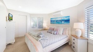 Une chambre à coucher comprenant un lit, une table de chevet, une lampe, une fenêtre avec des stores et un téléviseur fixé au mur.
