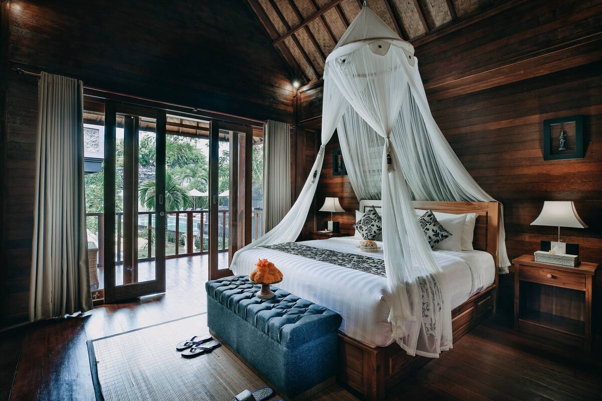 A bedroom with a canopy bed, wooden walls, a view through large windows, and a small bedside table with a lamp.
