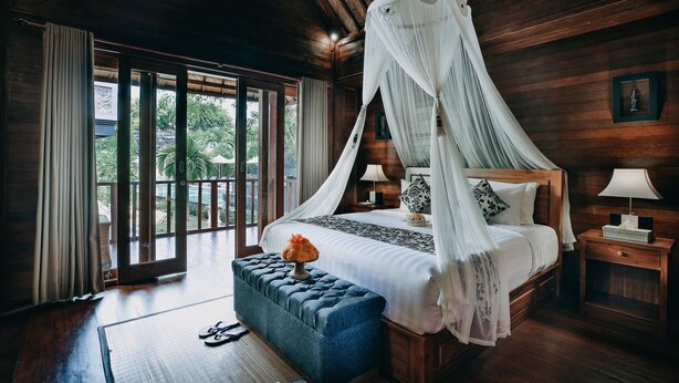 Un dormitorio con cama con dosel, paredes de madera, vistas a través de amplios ventanales y una pequeña mesita de noche con lámpara.