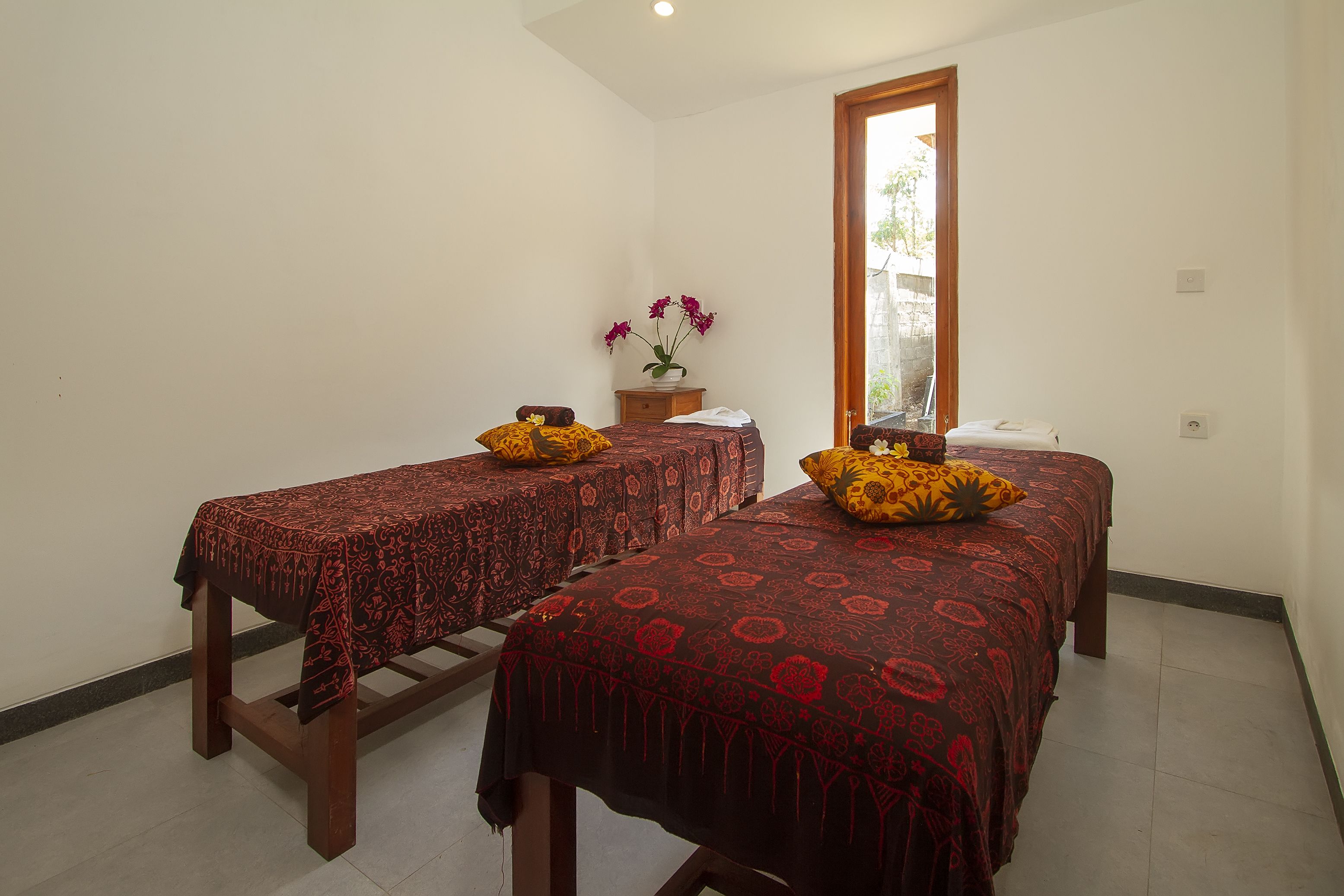 Une chambre avec deux lits simples, chacun agrémenté d’une couvre-lit et de coussins à motifs, une petite table de chevet en bois sur laquelle est posé un vase de fleurs, et une fenêtre donnant sur un paysage verdoyant.