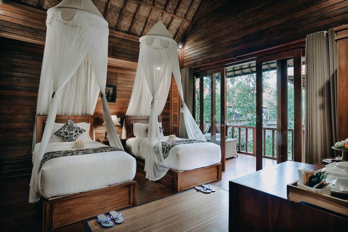 A room with two beds, each with a canopy, wooden floors, and large windows offering a view of greenery.