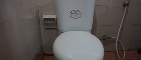 Un inodoro blanco con etiqueta Cotto, un accionador de carga y una manguera de bidé en un baño con baldosas.