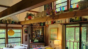 A cozy living room with a sofa, coffee table, and bookshelves filled with books. There is a dining table with chairs, and a large window with a view of greenery outside.
