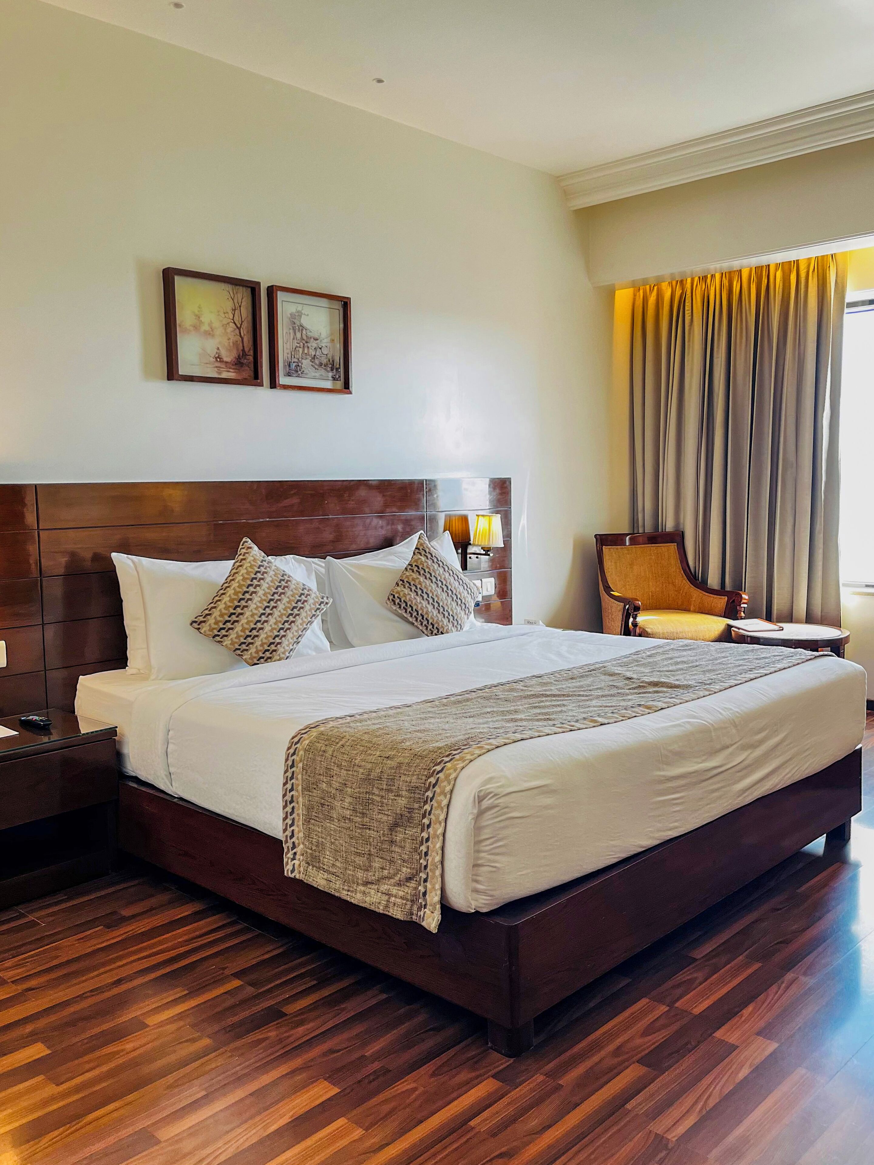 A hotel room with a large bed, wooden headboard, two bedside lamps, a chair, a small table, and a window with curtains.