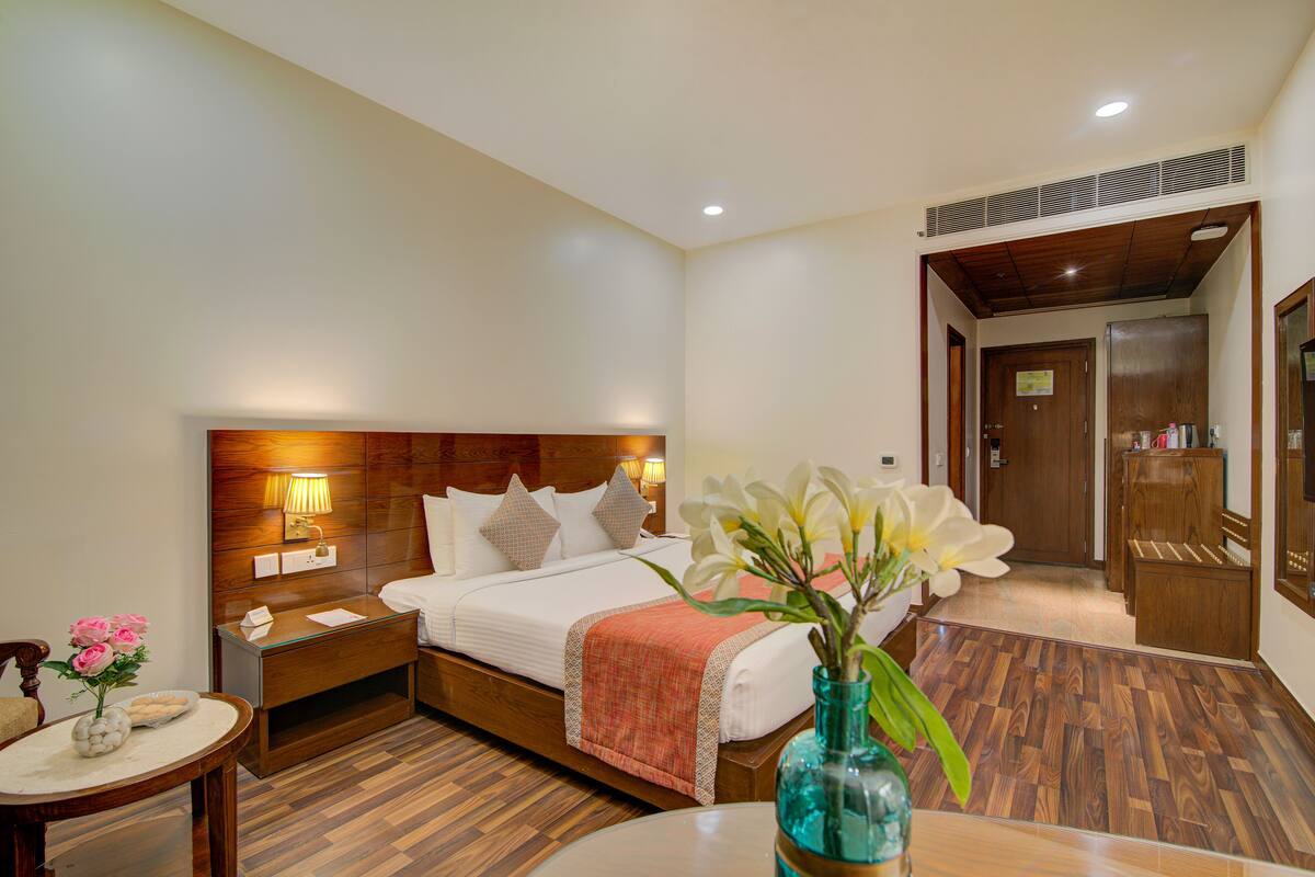 A hotel room with a wooden headboard, a bed with white linens, a nightstand with a lamp, a round table with a vase of flowers, and a wooden wardrobe.