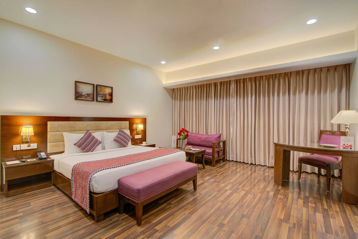 A hotel room with a large bed, bedside tables, a desk with a chair, and a view of the outside through curtains.