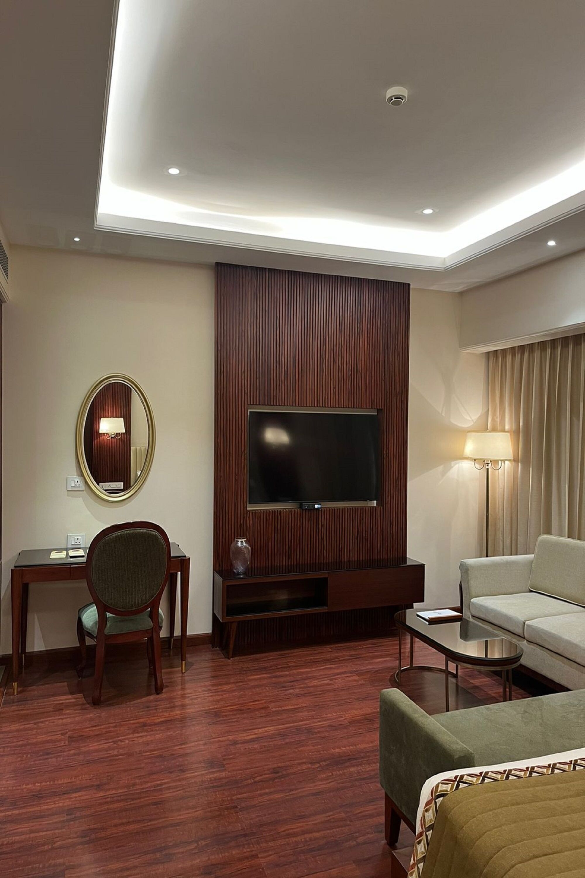 Une chambre d’hôtel dotée d’un téléviseur à écran plat fixé sur un mur en panneaux de bois, d’un bureau avec une chaise, d’un canapé et d’une petite table avec une lampe.