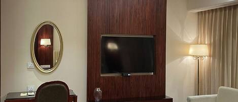 Habitación de hotel con una televisión de pantalla plana montada en una pared de paneles de madera, un escritorio con una silla, un sofá y una pequeña mesa con una lámpara.