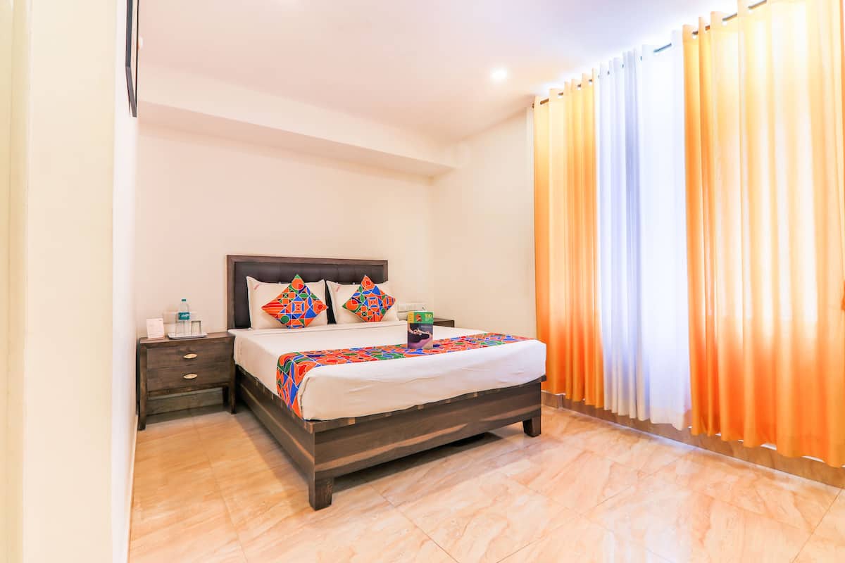 A neatly made bed with a patterned bedspread, a nightstand with a bottle and a box, and a window with orange curtains.