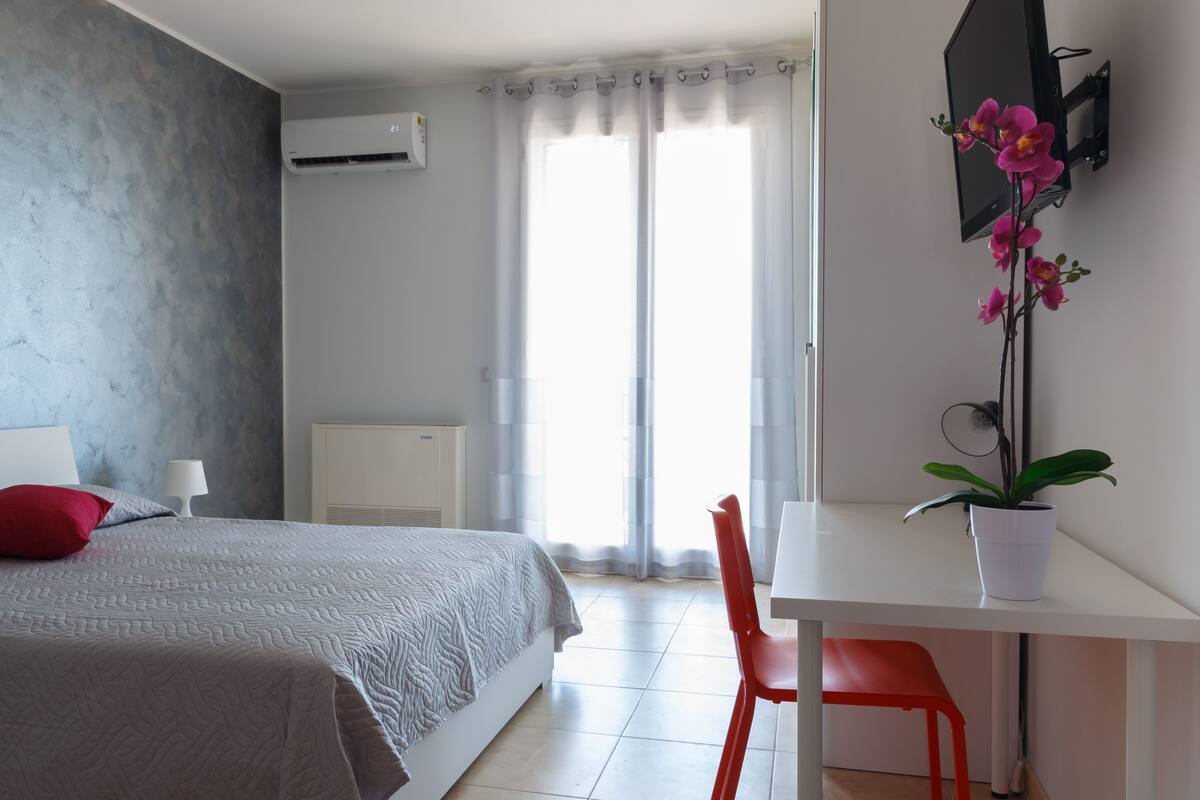 A bedroom with a bed, a desk, a chair, a potted plant, and a window with curtains.