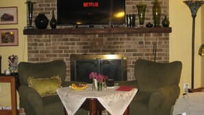 Una sala de estar con chimenea de ladrillo, un televisor mostrando 'NETFLIX', dos sillones, una mesita con mantel de encaje y objetos decorativos.