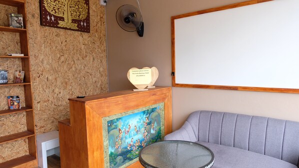 A reception area with a wooden desk, a glass table, a grey sofa, and a wall-mounted fan. - Frutta Hostel (Patong)