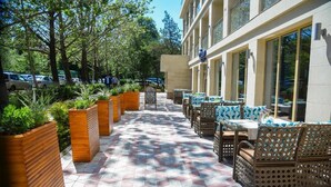 An outdoor patio with hexagonal patterned flooring, wooden planters, wicker furniture, and large windows on the building. - B Hotel Bishkek (Bishkek)