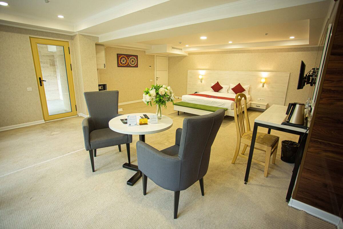 Ein Hotelzimmer mit einem Bett, zwei Stühlen, einem kleinen Tisch, einer Vase mit Blumen, einem Schreibtisch mit Lampe und einem Fernseher.