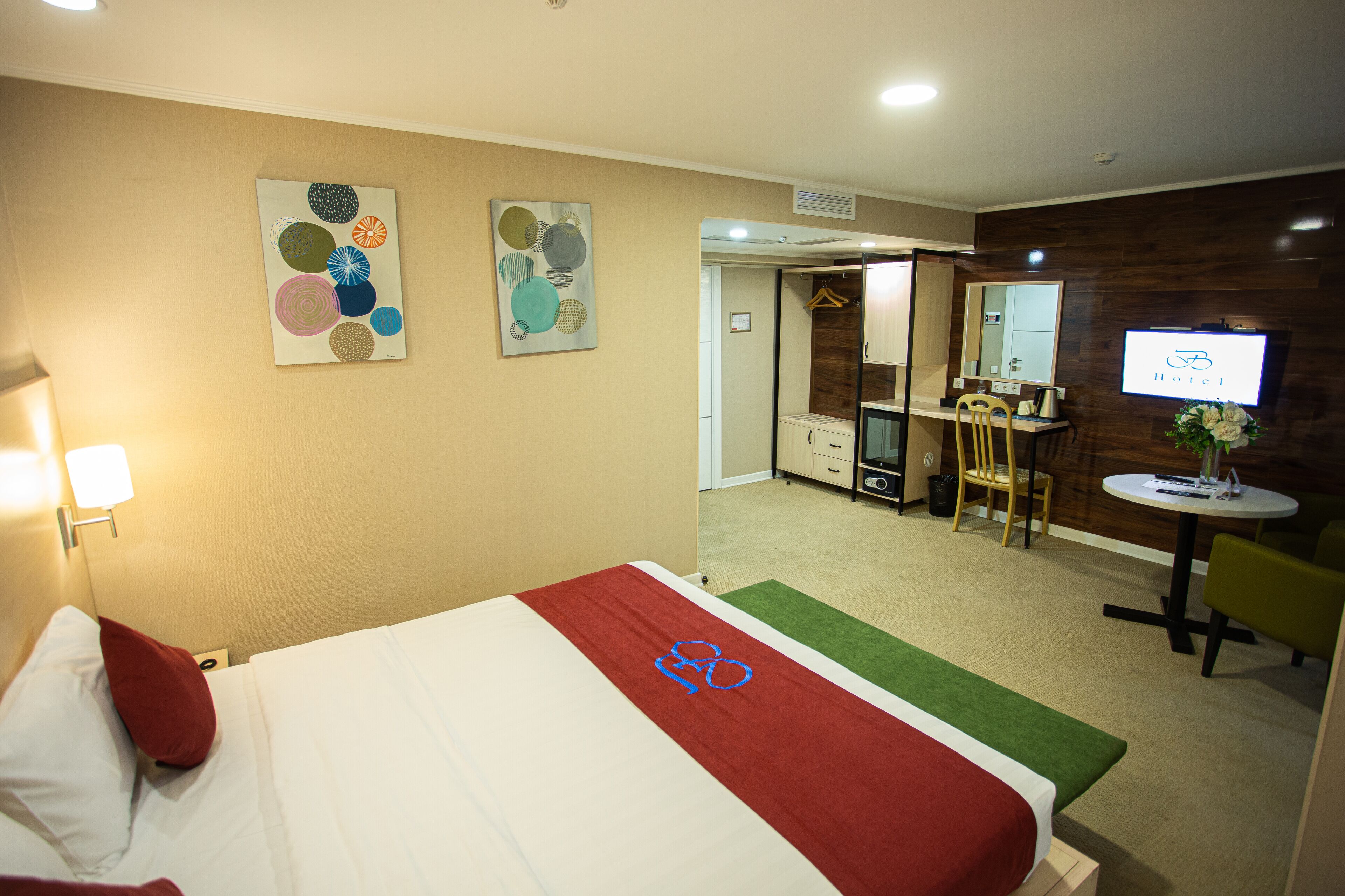 A hotel room with a bed, a desk, a chair, a TV, and artwork on the wall.
