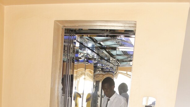 Interior de un ascensor con una persona frente al panel de control, una puerta de cristal y un piso de baldosas.