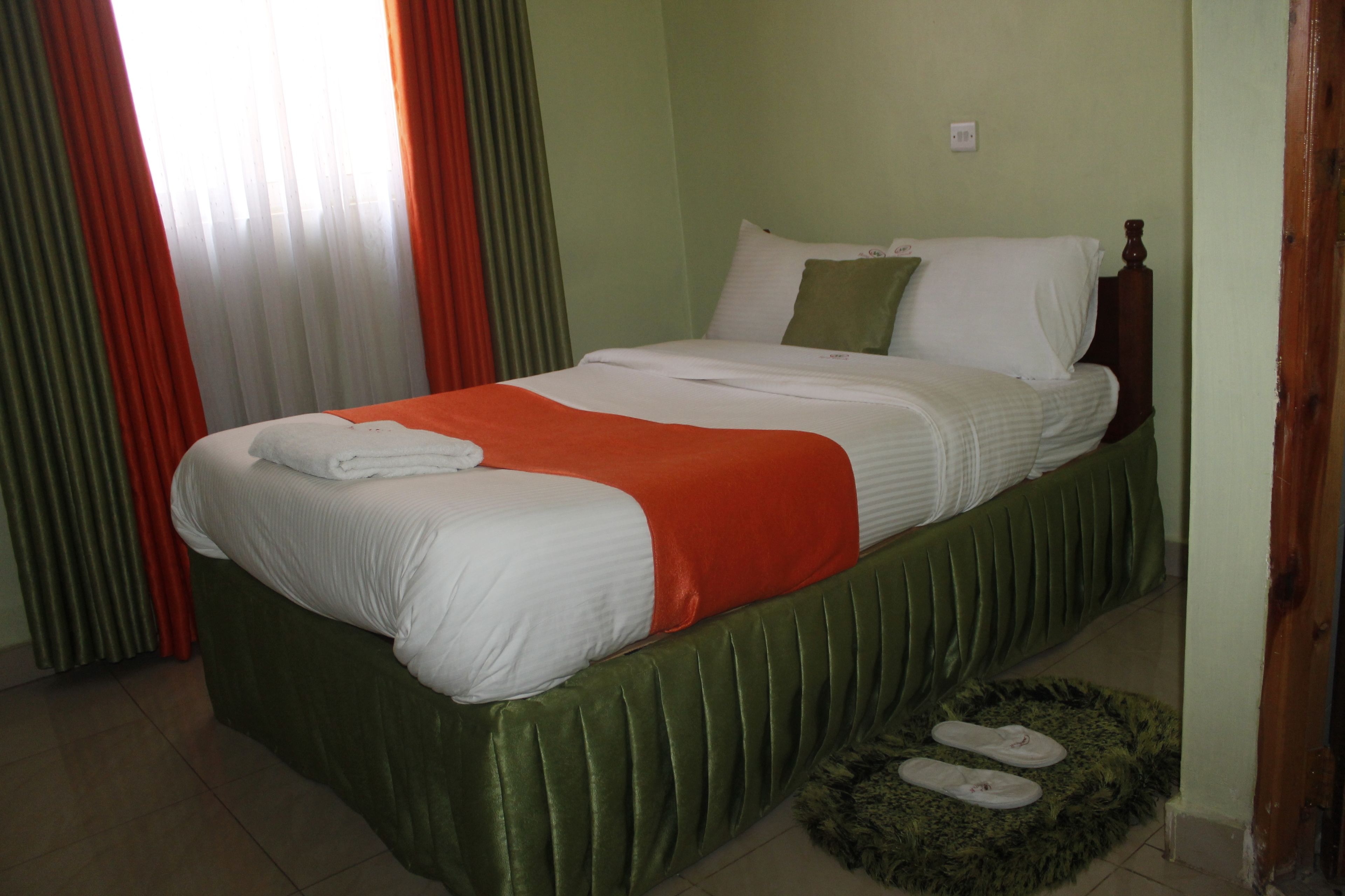 A neatly made bed with white and orange bedding, a green upholstered headboard, and a fluffy green rug with slippers.